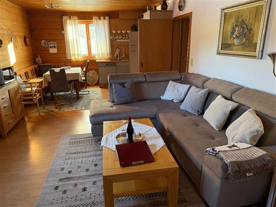 A cozy living room with a large gray couch and a wooden table. In the background, a dining table with chairs and a kitchen can be seen.