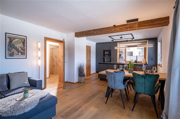 A modern living room with a cozy seating area and a dining area. The room has bright walls, wooden beams, and stylish furniture.