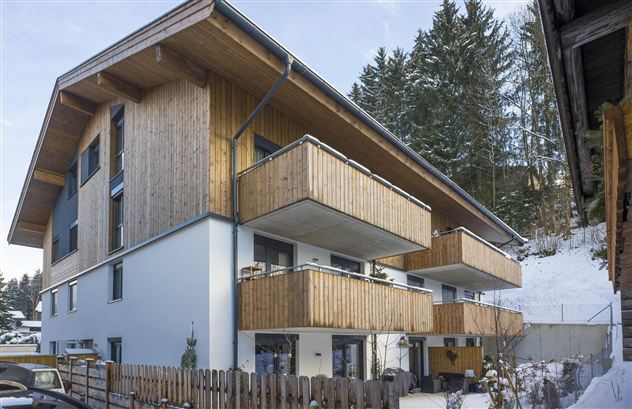 A modern house with wooden elements and several balconies. It stands in a snow-covered landscape, surrounded by trees.