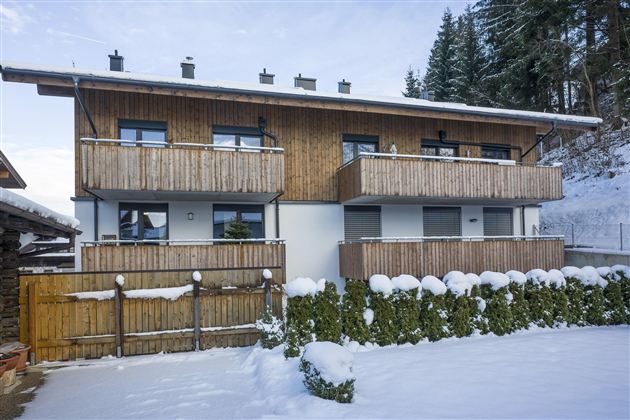 A modern two-story building with wooden siding in a snowy landscape. The garden is surrounded by a hedge and the sky is clear.