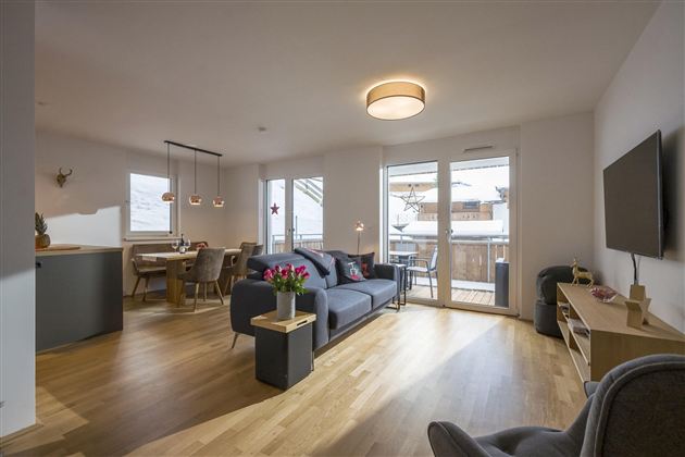 A modern living room with a comfortable sofa and a dining area. Large windows provide plenty of natural light and access to a balcony.