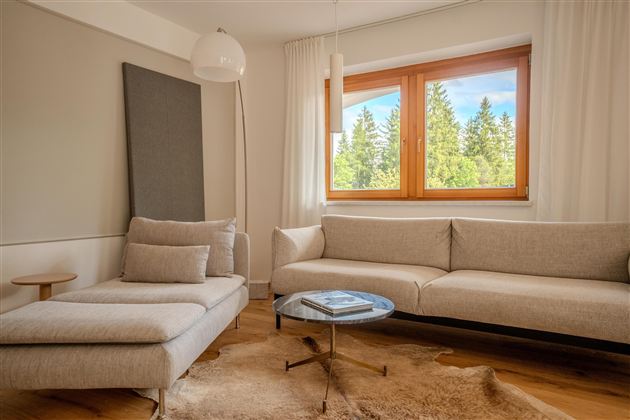 Ein modernes Wohnzimmer mit einem hellen Sofa und einem bequemen Sitzbereich. Durch das Fenster ist die grüne Landschaft sichtbar.