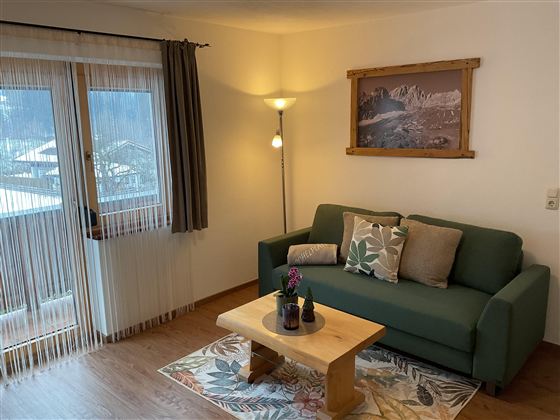 Ein einladendes Wohnzimmer mit einem grünen Sofa und einem Holztisch. Helles Licht strahlt durch die Fenster und schafft eine gemütliche Atmosphäre.