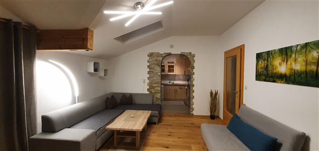 A modern living room with gray sofas and a wooden table. In the background, there is an open kitchen and a piece of art featuring a forest landscape on the wall.