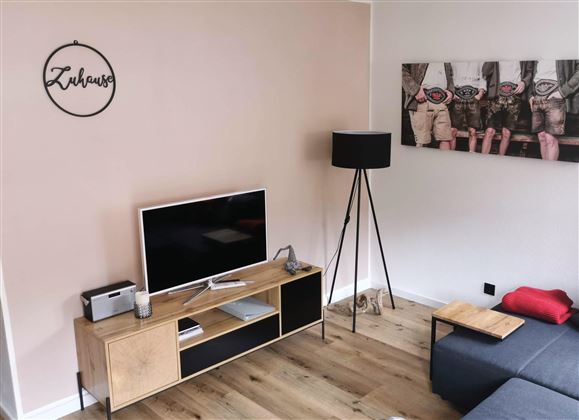 Ein modernes Wohnzimmer mit einem Fernseher auf einem Holzregal. Eine Stehlampe und ein Wandbild ergänzen die gemütliche Atmosphäre.