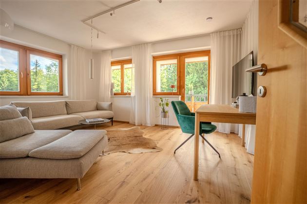 Ein helles Wohnzimmer mit großen Fenstern und einem modernen Sofa. Ein Schreibtisch mit einem Stuhl steht ebenfalls im Raum und sorgt für eine einladende Atmosphäre.