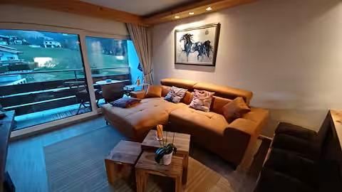 Een gezellige woonkamer met een bruine bank en een houten tafel. Grote ramen bieden uitzicht op het omliggende landschap.