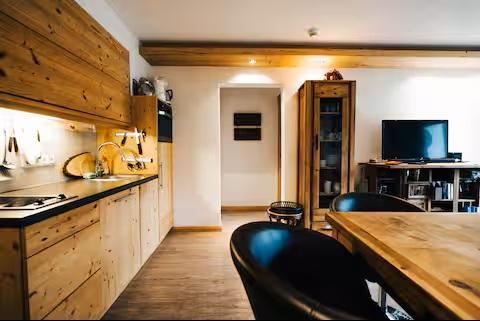 Een moderne keuken met houten meubels en een eettafel. Op de achtergrond staat een tv en er is een open ruimte.