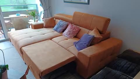 Een gezellige, oranje bank met kleurrijke kussens staat in een lichte kamer. Op de achtergrond zijn ramen en een tafel zichtbaar.