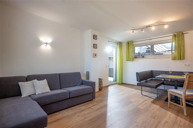 A modern living room with a gray couch and a table for four people. Large windows with green curtains let in plenty of light.