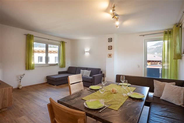 A cozy living room with a seating area and a dining table. The walls are light, and green curtains create a friendly atmosphere.