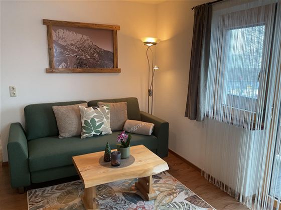 Ein gemütlicher Wohnbereich mit einem grünen Sofa und einem Holztisch. An der Wand hängt ein Bild von Bergen, und das Fenster lässt viel Licht herein.