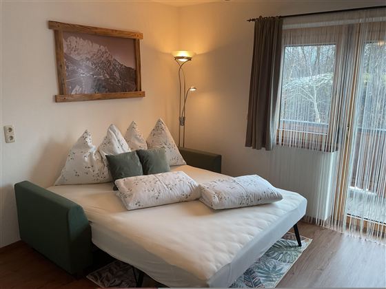 Ein gemütliches Schlafzimmer mit einem Bett und mehreren Kissen. Der Raum hat große Fenster und eine angenehme Atmosphäre.