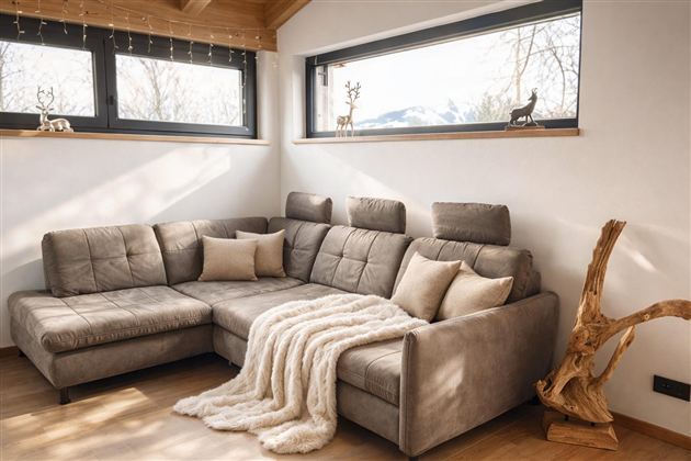 Ein modernes Wohnzimmer mit einem beigen Sofa und einer gemütlichen Decke. Große Fenster bieten einen Blick auf die Natur draußen.