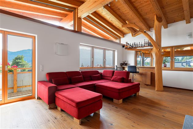 Ein modernes Wohnzimmer mit rotem Sofa und Holzbalkendecke. Große Fenster bieten einen Blick auf die umliegende Landschaft.<br/>Mit einer gemütlichen Bar integriert.