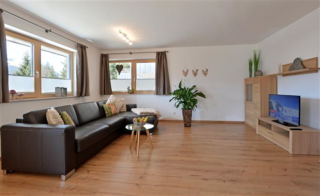 Ein modernes Wohnzimmer mit einem schwarzen Sofa und einem Holztisch. Große Fenster lassen viel Licht herein und es gibt Pflanzen zur Dekoration.