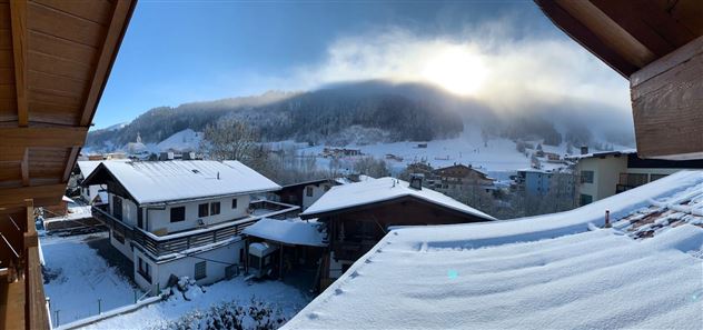A picturesque winter scene with snow-covered roofs and mountains in the background. The sun shines through the clouds, giving the landscape a warm light.