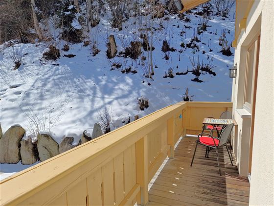 A wooden balcony with a table and two chairs overlooks a snow-covered landscape. The sun shines on the snowy area in the background.