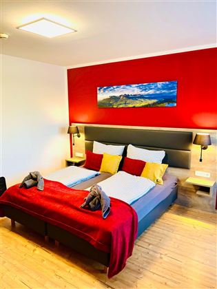 Een moderne slaapkamer met een groot dubbelbed en kleurige kussens. Aan de muur hangt een landschapsschilderij, en de ruimte heeft een gezellige sfeer.