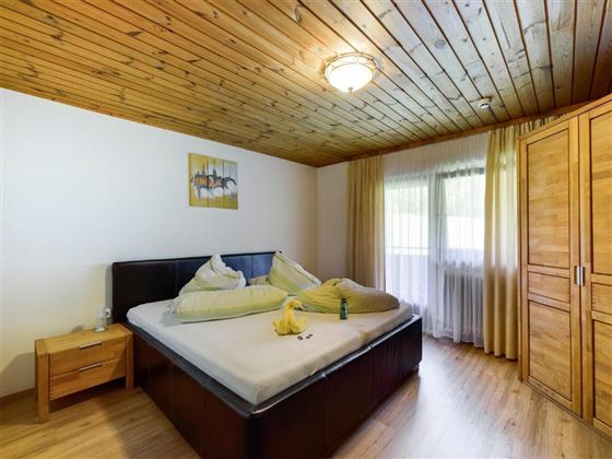 A cozy bedroom with a large bed and a wooden ceiling. Natural light flows through the windows, which are framed by light curtains.