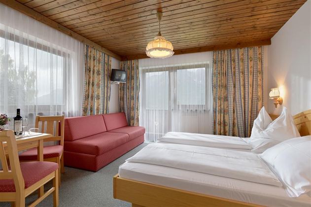 A cozy hotel room with two single beds and a red sofa. The room has large windows and a bright, inviting atmosphere.