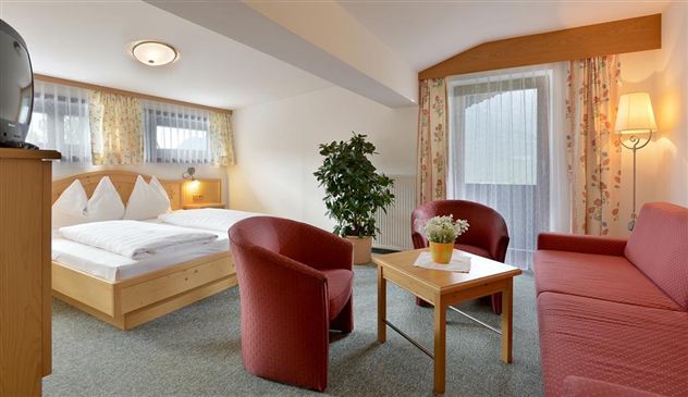 A cozy room with a double bed, red armchairs, and a table. Bright light comes through the windows, and a plant adds to the pleasant atmosphere.