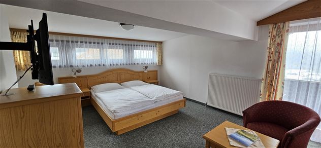 Een lichte slaapkamer met een tweepersoonsbed en houten meubels. Een televisie en een comfortabele fauteuil zijn ook aanwezig.
