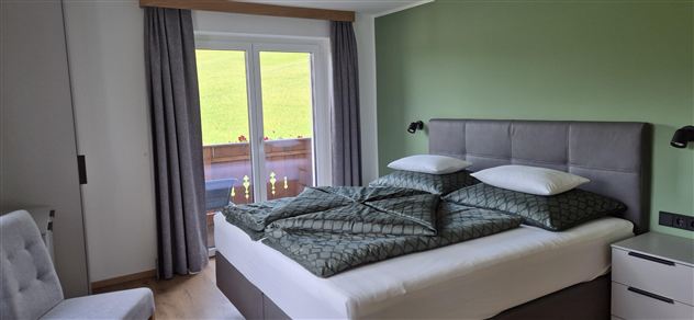 A modern bedroom with a large bed and green walls. Large windows let in plenty of daylight and offer a view of the outdoors.