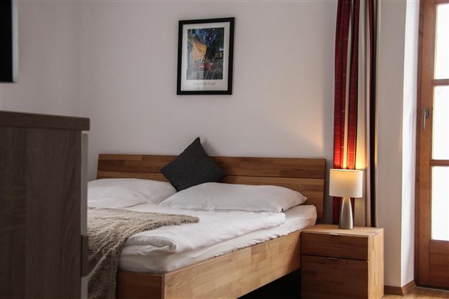 Ein gemütliches Schlafzimmer mit einem Doppelbett und künstlerischer Wanddekoration. Es gibt einen Nachttisch mit einer Lampe und große Fenster mit Vorhängen.