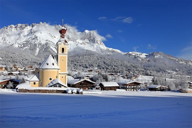 A picturesque winter landscape with snow-covered hills and charming cottages. In the foreground is a yellow church against a clear blue sky.
