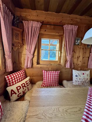 A cozy wooden room with checkered curtains and cushions. A window at the table offers a view outside.