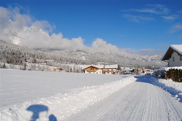 A snowy landscape with a clear blue sky. In the background, houses and mountains can be seen.