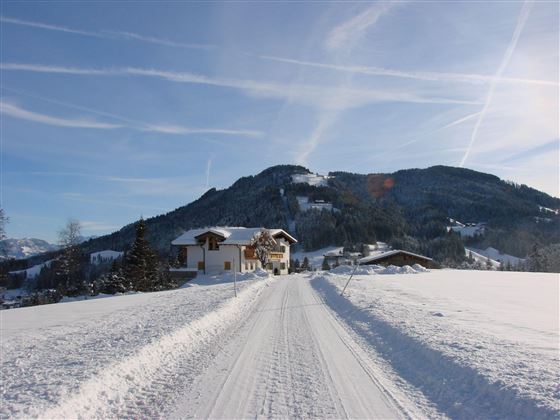 Eine verschneite Landschaft mit einem Wohnhaus und einer ruhigen Straße. Im Hintergrund sind sanfte Berge und ein klarer Himmel zu sehen.
