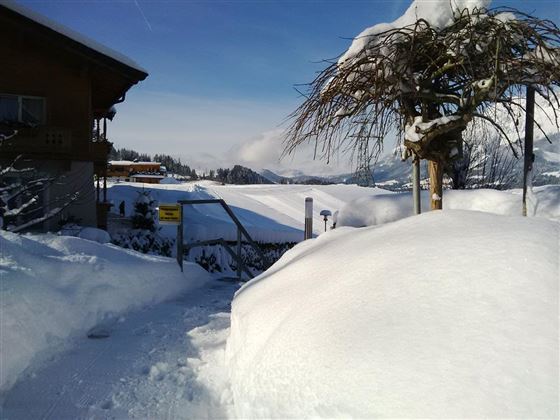 Eine schneebedeckte Landschaft mit einem typischen Holzhaus im Hintergrund. Die Szene vermittelt eine ruhige Winteratmosphäre mit klarem Himmel und Bergen in der Ferne.