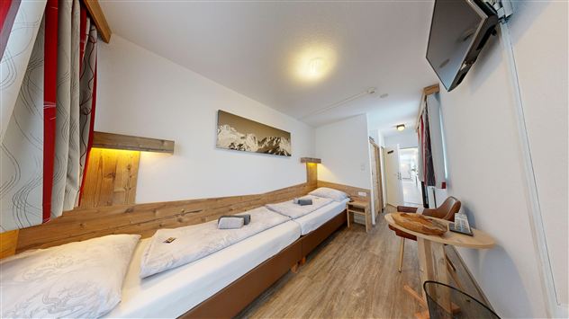 A modern room with two single beds, wooden details, and a television. The walls are light, and there is a small table as well as a chair.